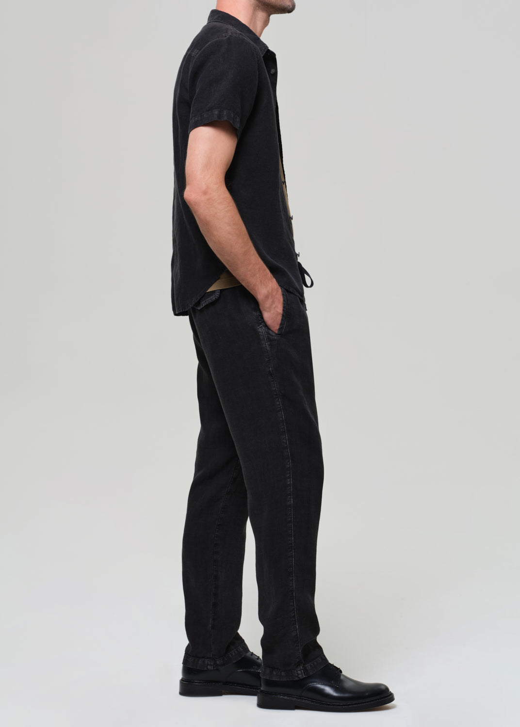 Person wearing a black short-sleeve linen shirt over a beige t-shirt, paired with black linen pants on a grey background