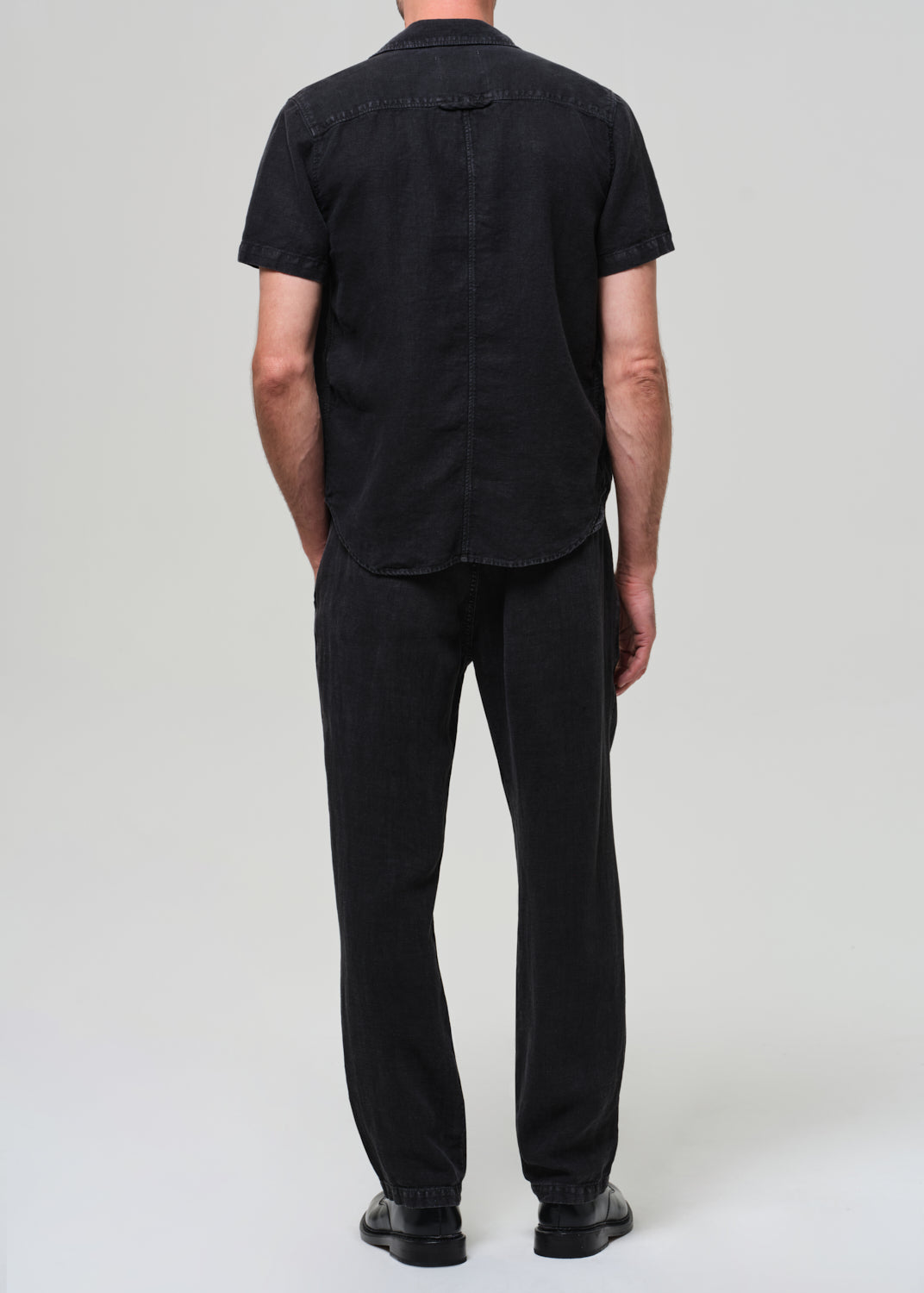 Person wearing a black short-sleeve linen shirt over a beige t-shirt, paired with black linen pants on a grey background