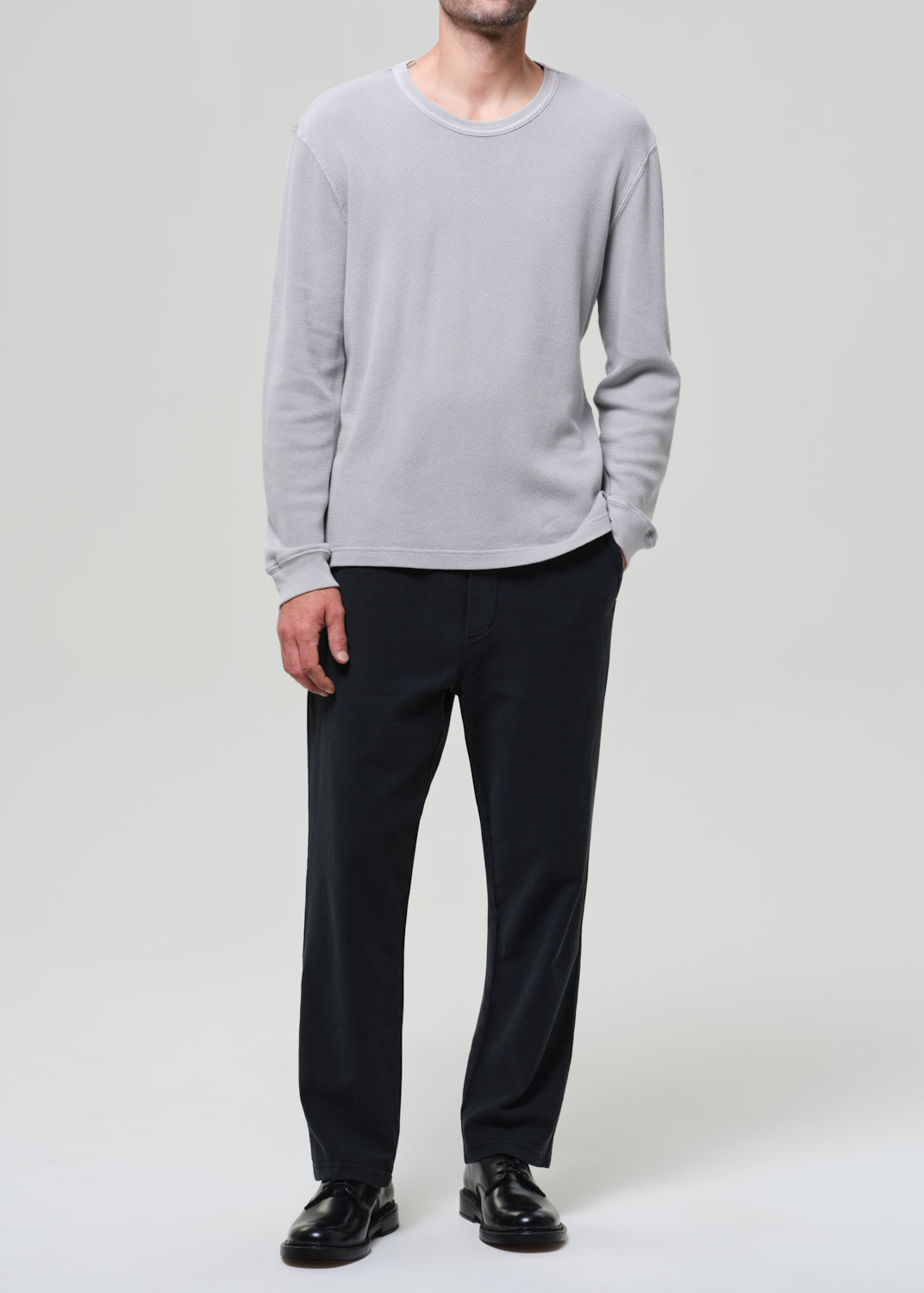 Person wearing a light gray crewneck and charcoal pants on a grey background