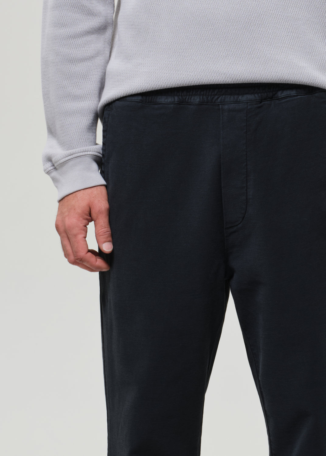 Person wearing a light gray crewneck and charcoal pants on a grey background