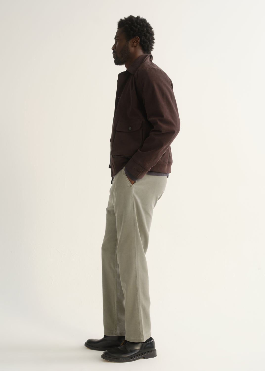 Person wearing light beige straight leg pants with a dark brown jacket and black shoes, side full body view on a light grey background.