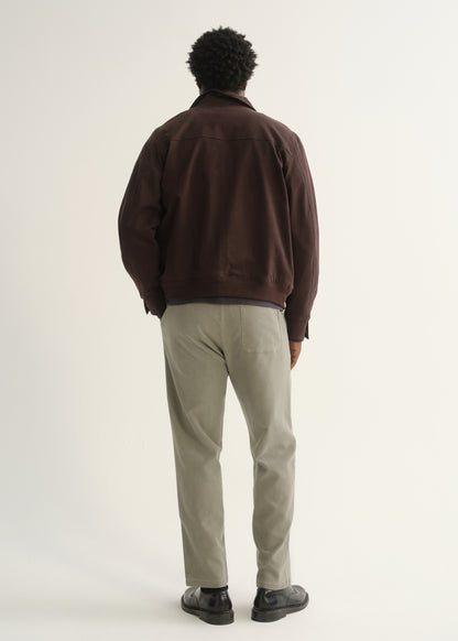Person wearing light beige straight leg pants with a dark brown jacket and black shoes, back full body view on a light grey background.