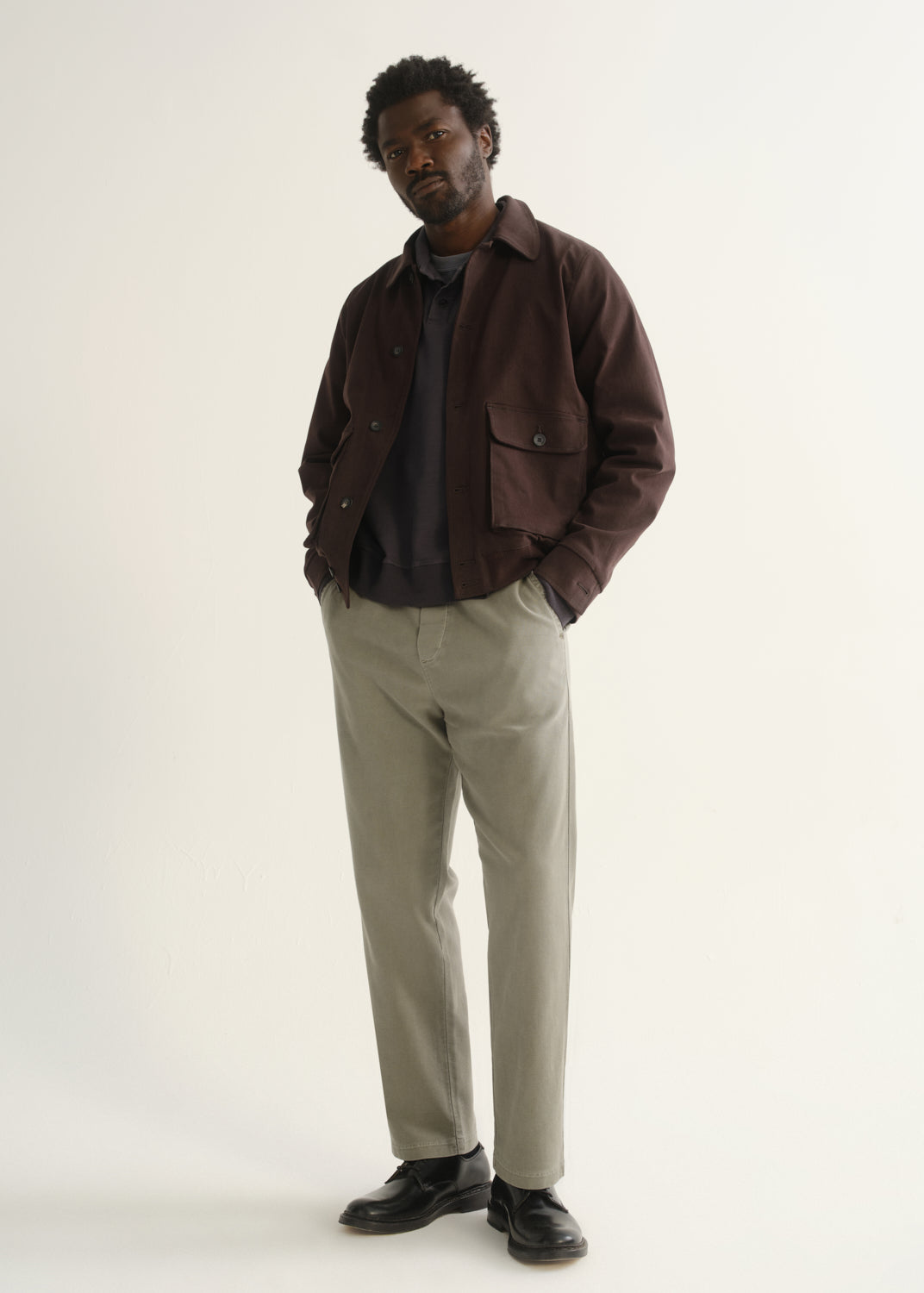 Person wearing light beige straight leg pants with a dark brown jacket, dark shirt, and black shoes, angled front full body view on a light grey background.