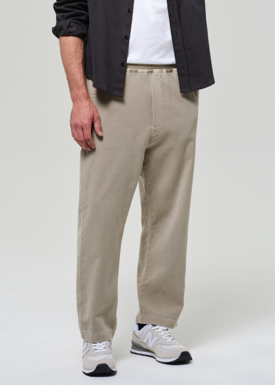 Person wearing a grey shirt, white t-shirt, beige pants, and white sneakers on a plain background