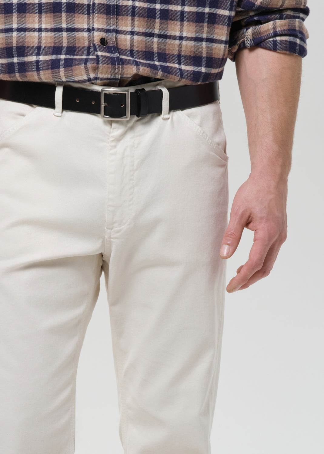 A close-up of a person wearing off-white chino pants with a black belt and a blue, brown, and white plaid shirt, shown from the waist down against a grey background.