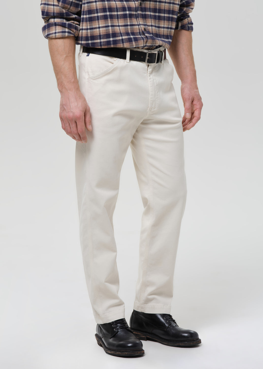 A person wearing off-white straight-leg chinos, a navy and tan plaid shirt, a black belt, and black leather shoes, standing against a grey background.