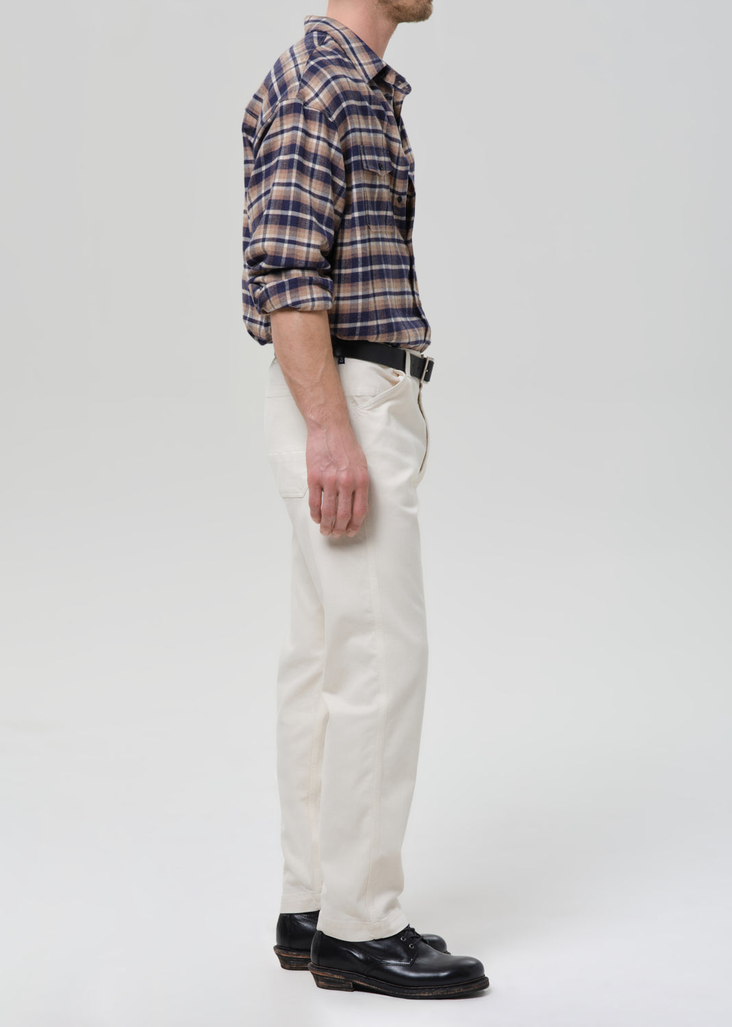 A man shown in profile wearing a navy and tan plaid button-down shirt, off-white trousers, a black belt, and black leather shoes, photographed against a grey background.