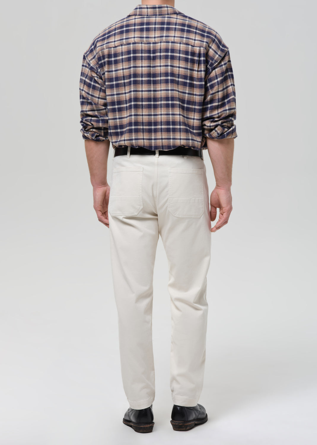 A man viewed from the back wearing a navy, beige, and brown plaid long-sleeve button-down shirt, off-white chino pants, a black belt, and black shoes, against a grey background.