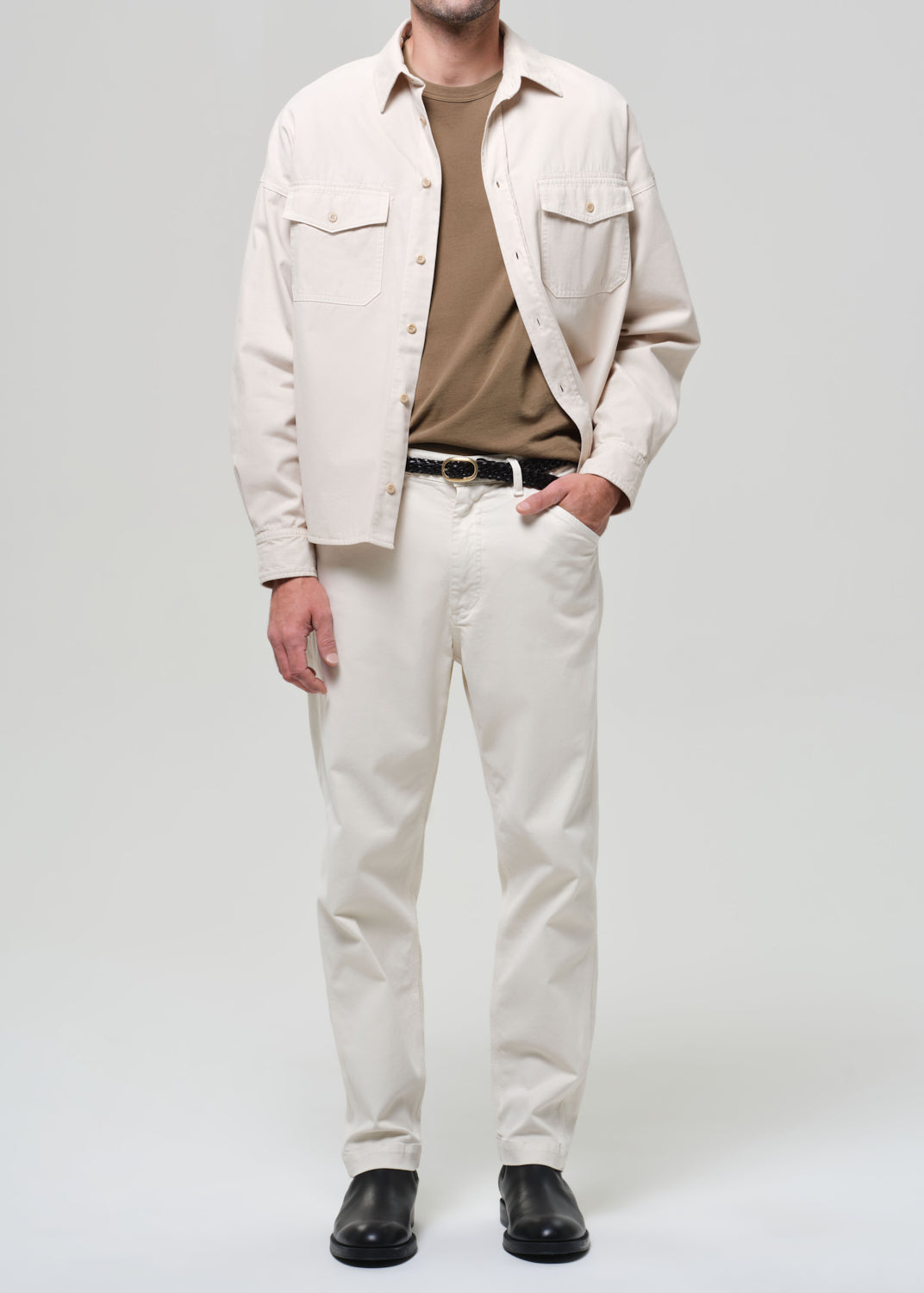 Man wearing a beige shirt, brown t-shirt, white pants, and black shoes on a light gray background