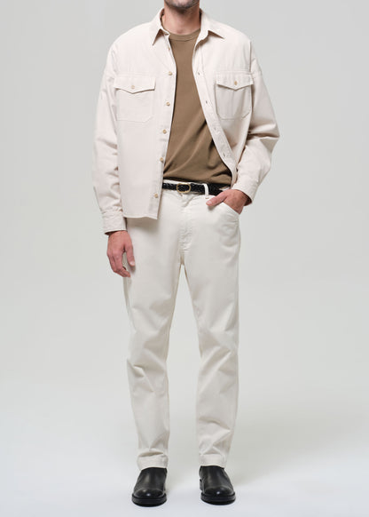 Man wearing a beige shirt, brown t-shirt, white pants, and black shoes on a light gray background