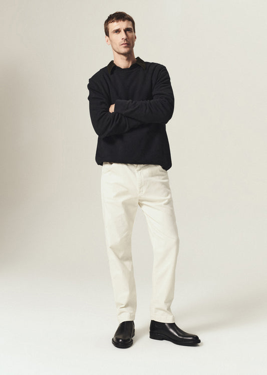 A man modeling a black long-sleeve knit top and off-white trousers, standing with arms crossed against a plain light beige studio background.