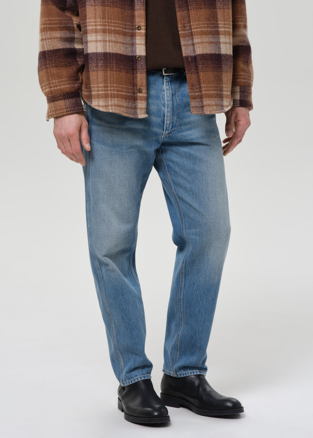 Person wearing light blue straight leg jeans with a brown plaid overshirt on a grey background.