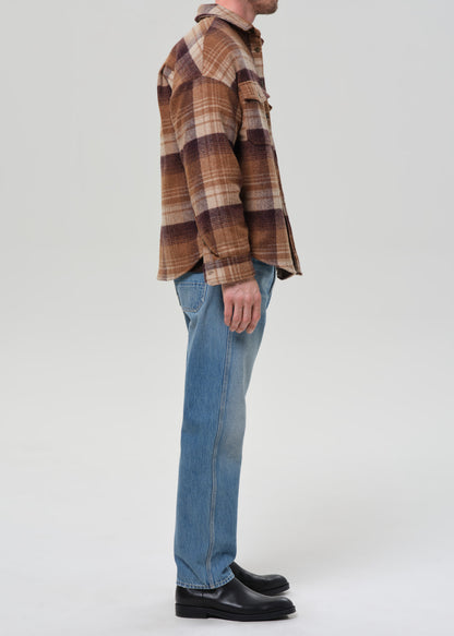 Person wearing light blue straight leg jeans with a brown plaid overshirt viewed from the side on a grey background.