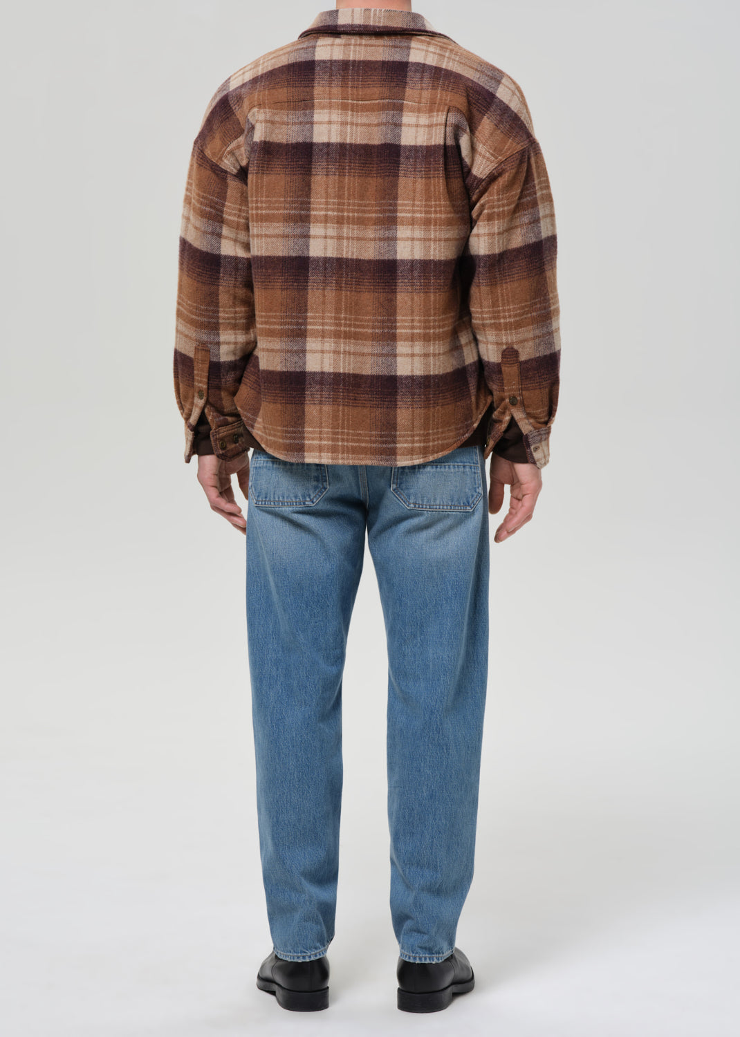 Person wearing light blue straight leg jeans with a brown plaid overshirt viewed from the back on a grey background.