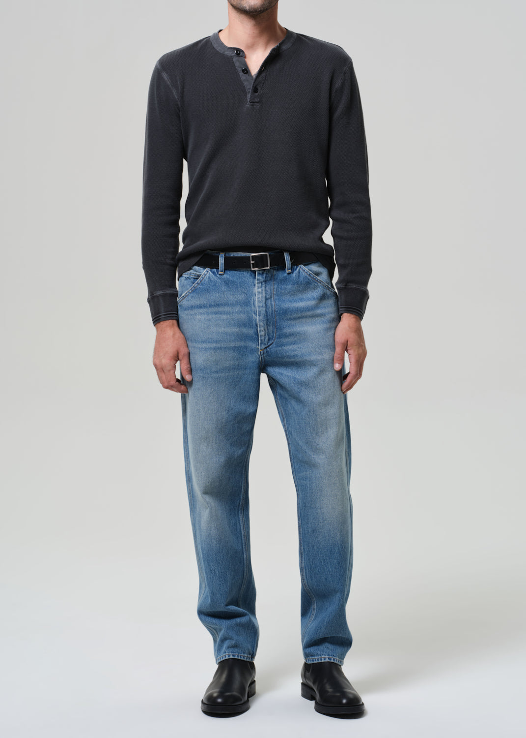 Person wearing a dark long-sleeve shirt, blue jeans, and black shoes on a grey background