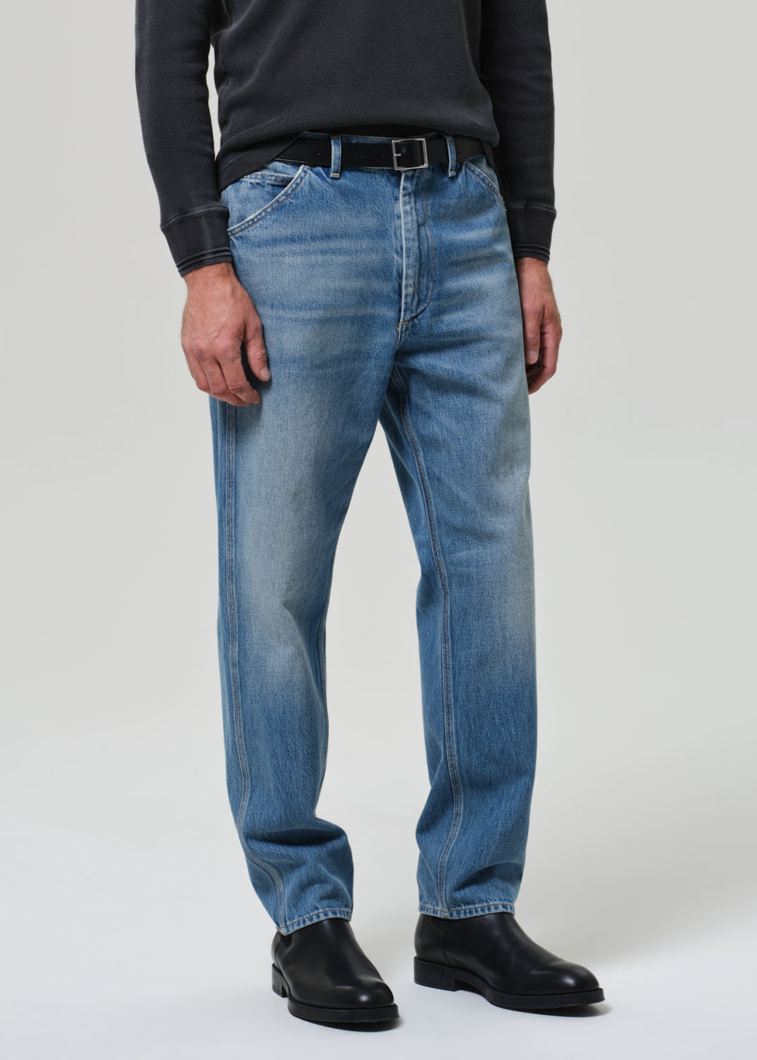 Person wearing a dark long-sleeve shirt, blue jeans, and black shoes on a grey background