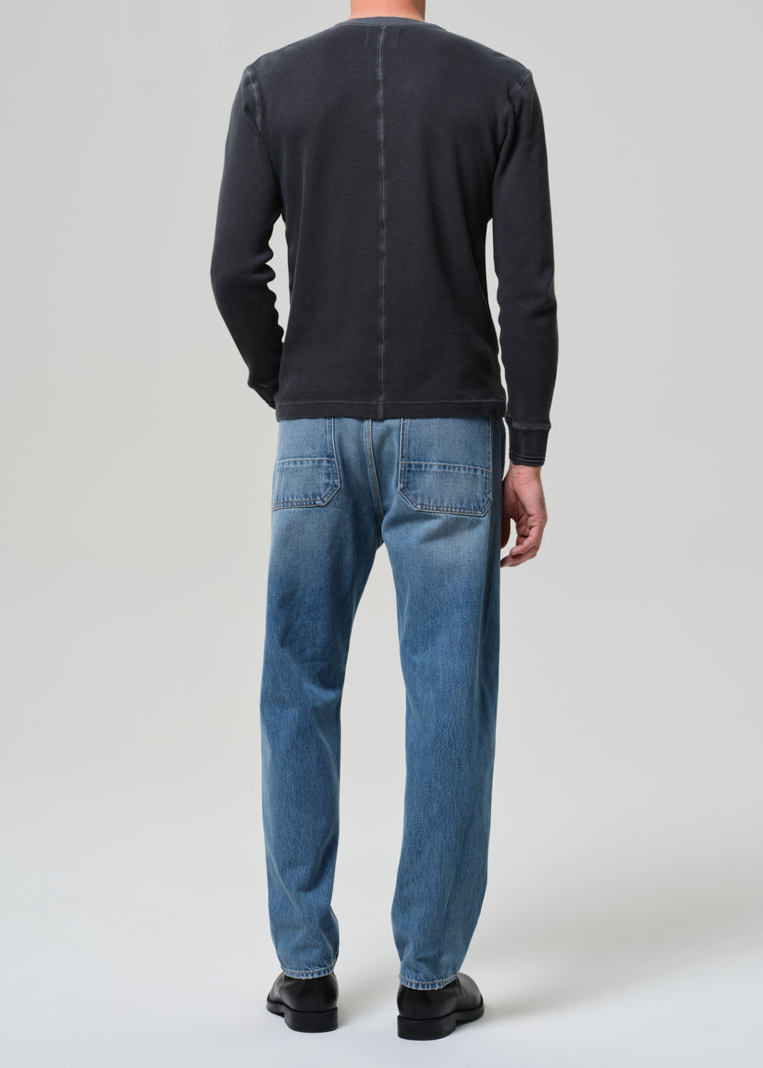 Person wearing a dark long-sleeve shirt, blue jeans, and black shoes on a grey background