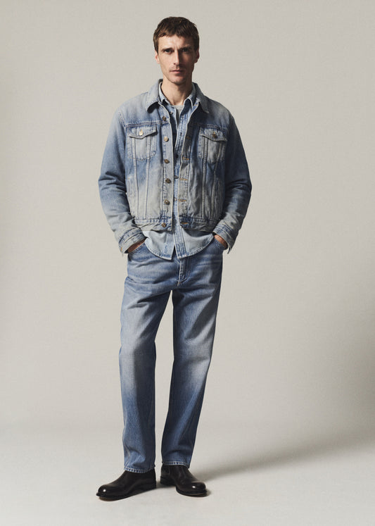 A man modeling a light-wash blue denim jacket and matching blue jeans with black shoes, standing against a plain light gray studio background.