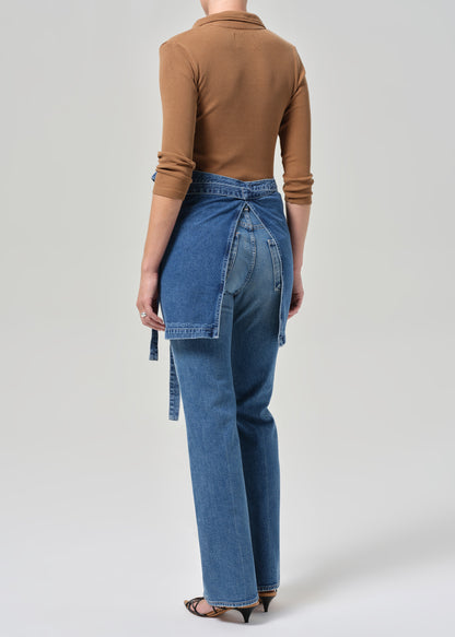 Person wearing a brown top and blue apron and jeans with a white background