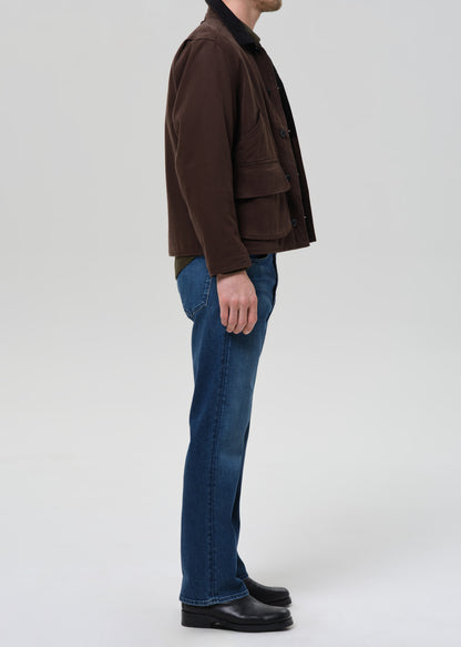 Person wearing a brown jacket and blue jeans on a grey background