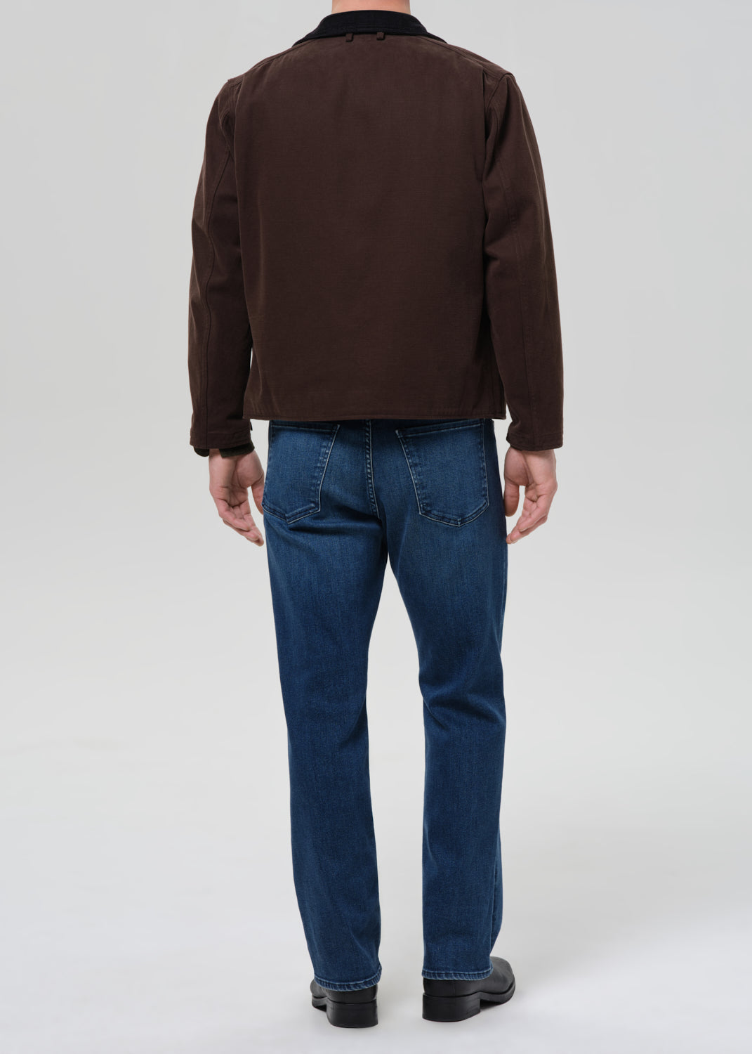 Person wearing a brown jacket and blue jeans on a grey background