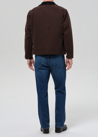 Person wearing a brown jacket and blue jeans on a grey background