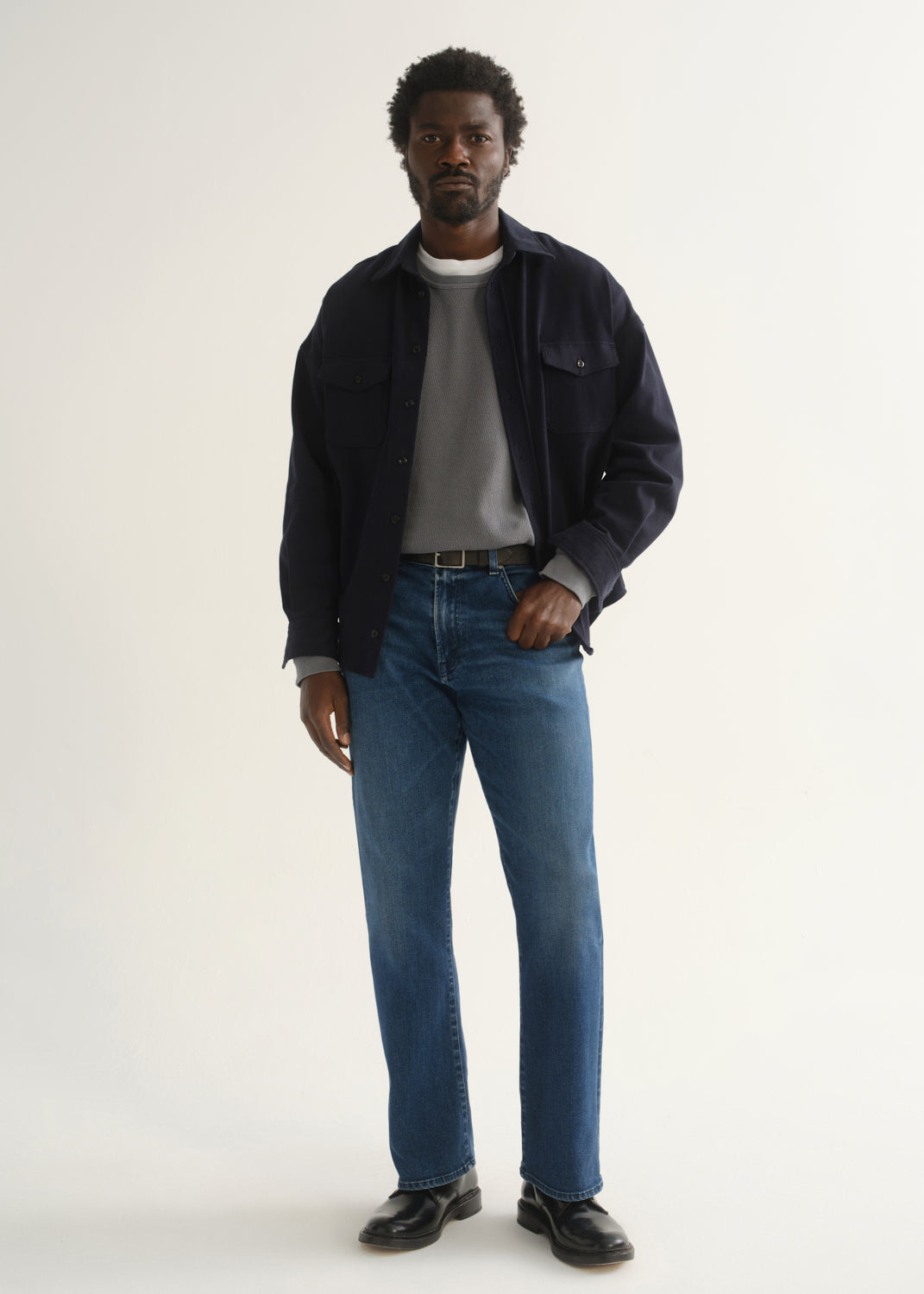 Person wearing medium blue straight leg jeans with a navy jacket, grey t-shirt, and black shoes, front full body view on a light grey background.