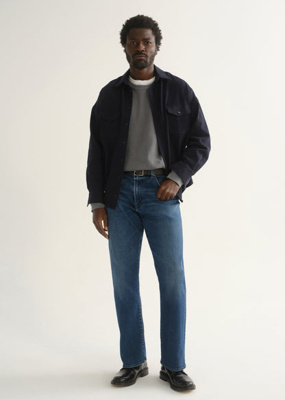 Person wearing medium blue straight leg jeans with a navy jacket, grey t-shirt, and black shoes, front full body view on a light grey background.