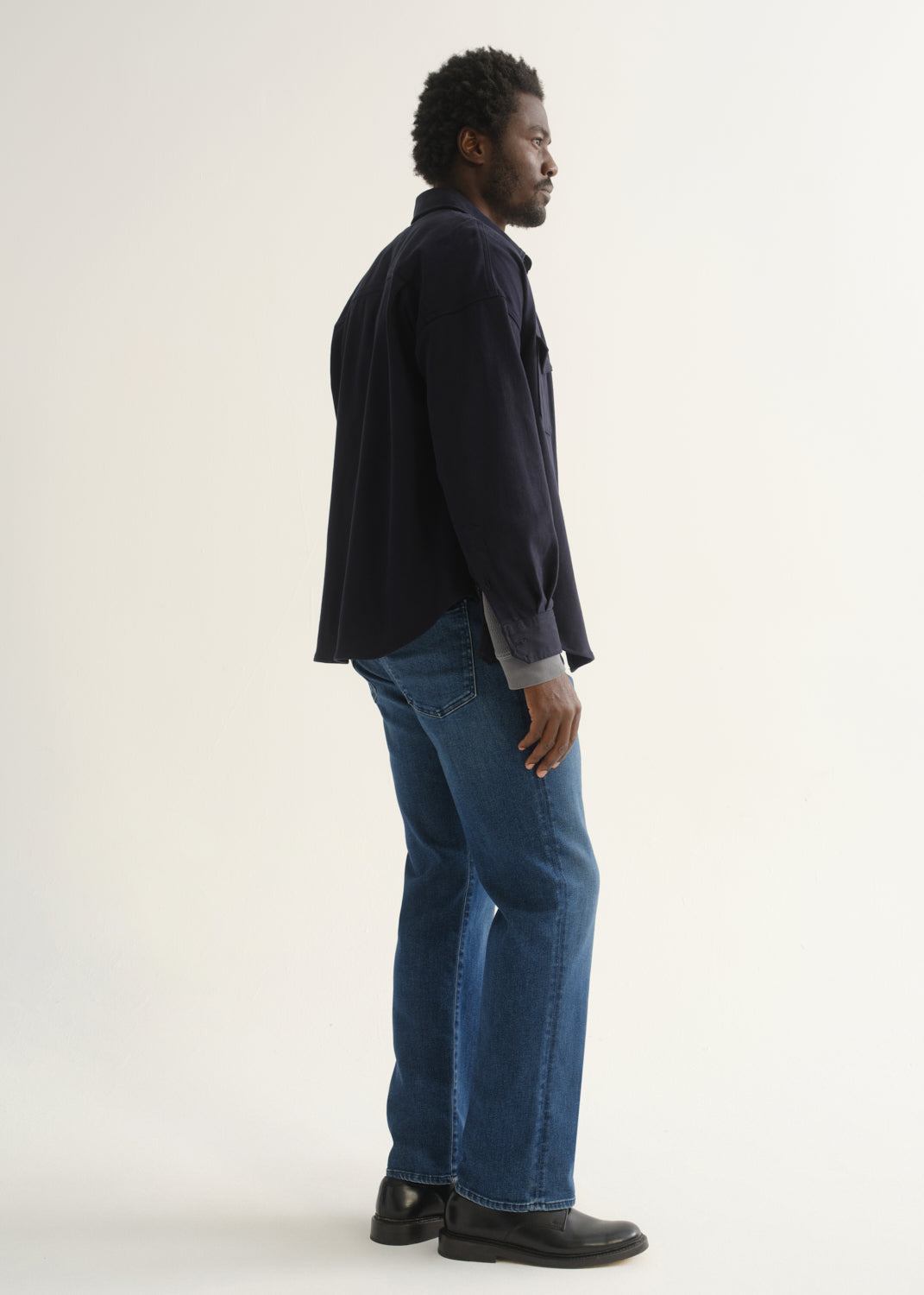 Person wearing medium blue straight leg jeans with a navy jacket and black shoes, side full body view on a light grey background.