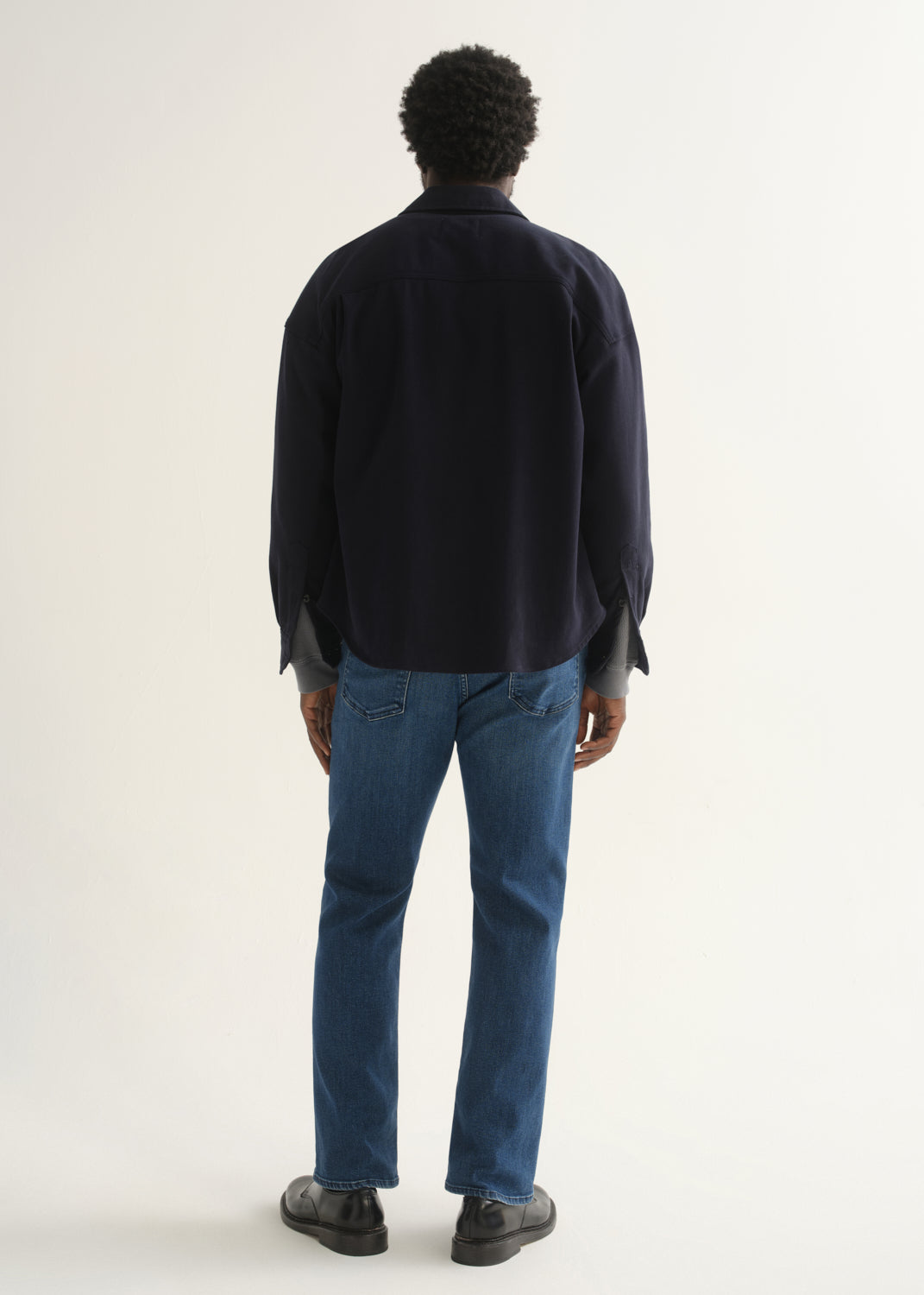 Person wearing medium blue straight leg jeans with a navy jacket and black shoes, back full body view on a light grey background.