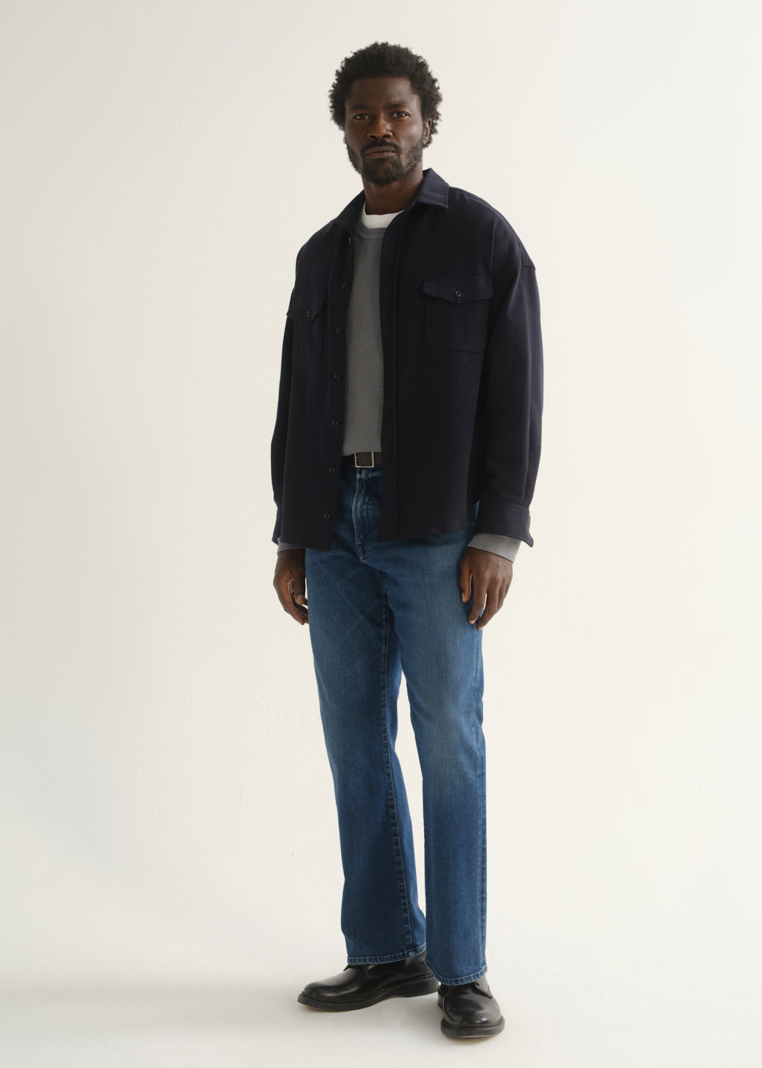 Person wearing medium blue straight leg jeans with a navy jacket, grey t-shirt, and black shoes, angled front full body view on a light grey background.