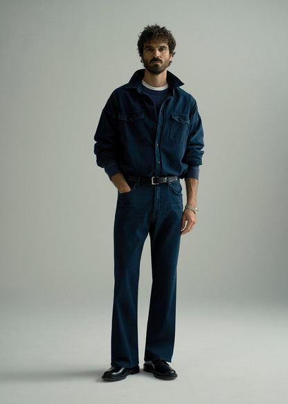 Man wearing dark indigo straight leg jeans with a matching dark indigo denim shirt layered over a navy crewneck top on a grey background.