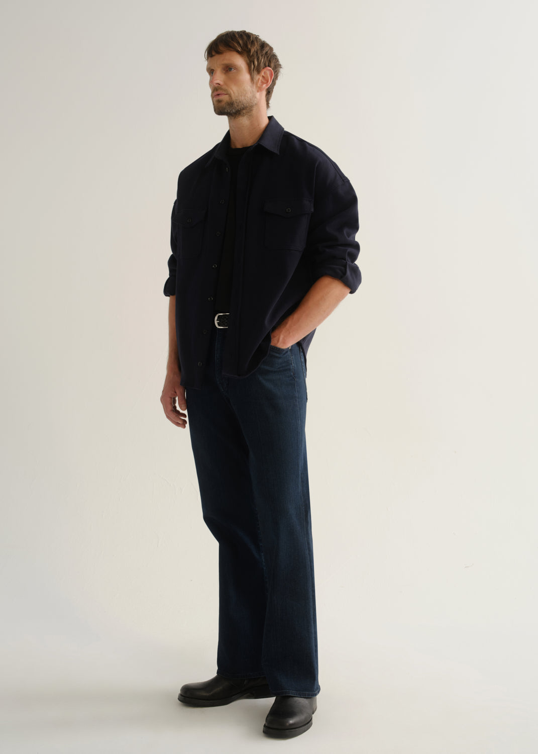 Person wearing dark indigo wide leg jeans with a navy long sleeve shirt and black boots, angled front full body view on a light grey background.