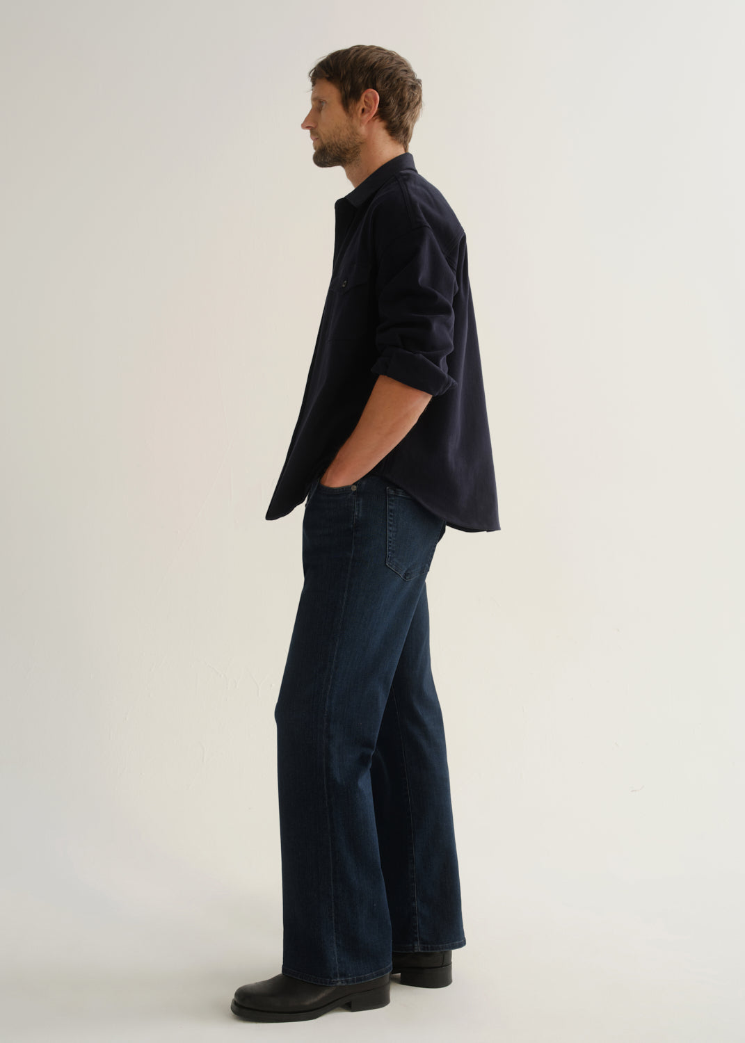 Person wearing dark indigo wide leg jeans with a navy long sleeve shirt and black boots, side full body view on a light grey background.