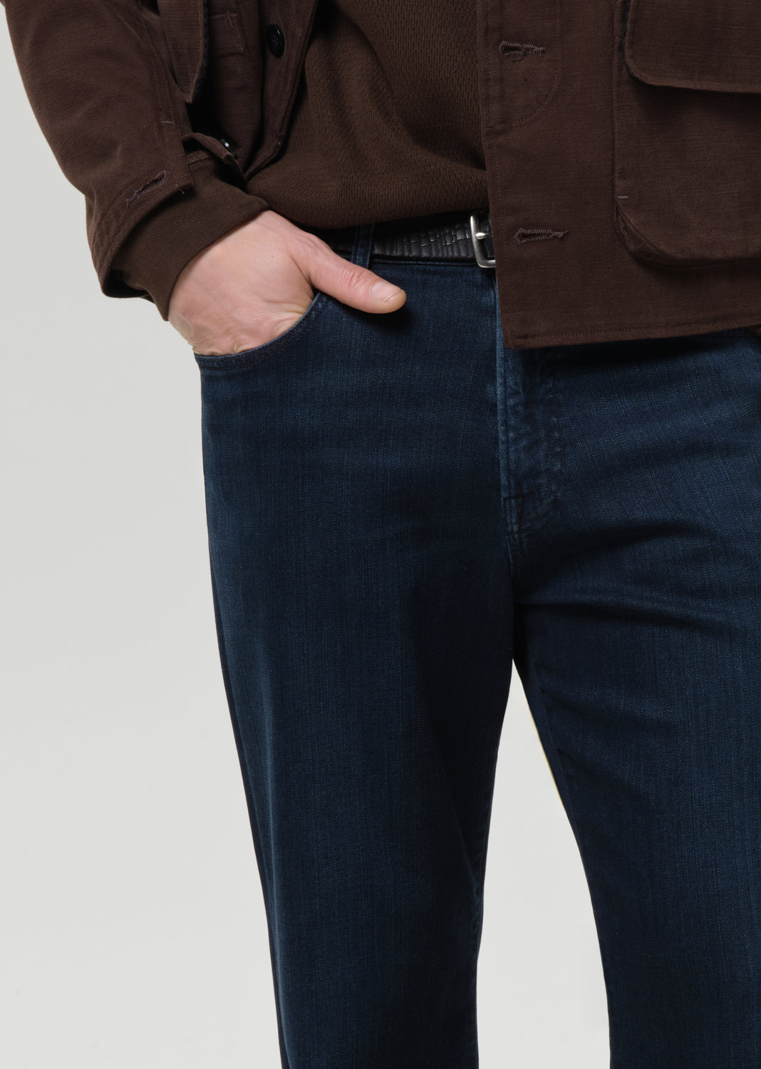 Person wearing a brown jacket and dark blue jeans on a grey background.
