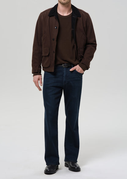 Man wearing a brown jacket, brown shirt, and dark blue jeans on a grey background.