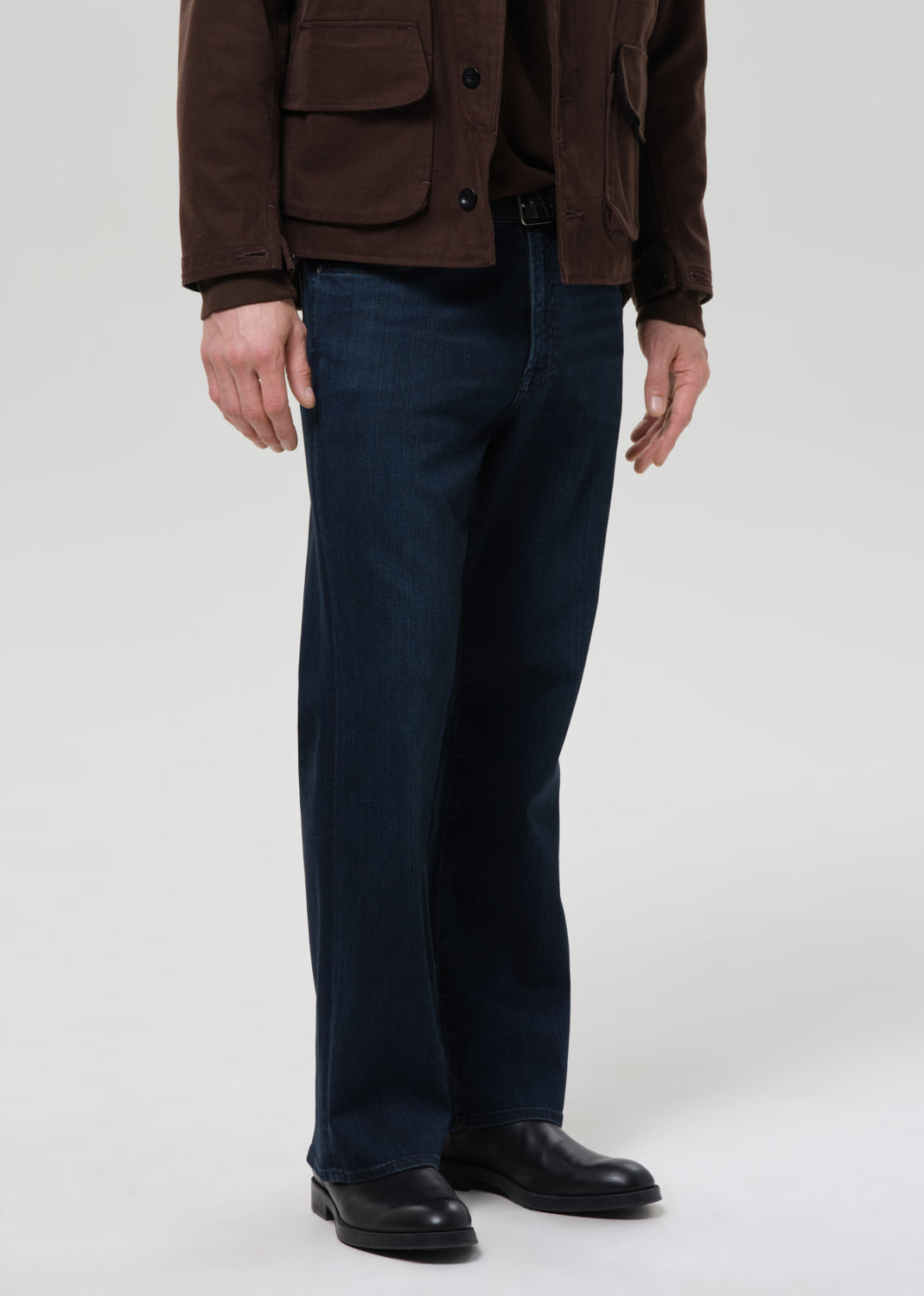 Person wearing a brown jacket, blue pants, and black shoes on a grey background.