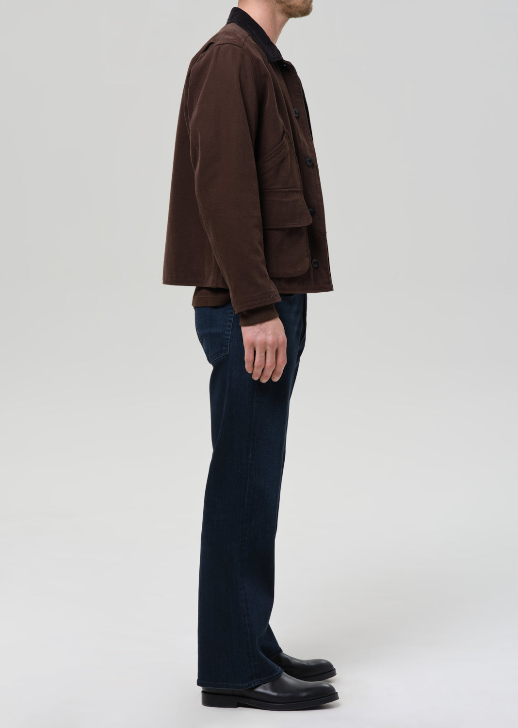 Person wearing a brown jacket and dark pants on a grey background.