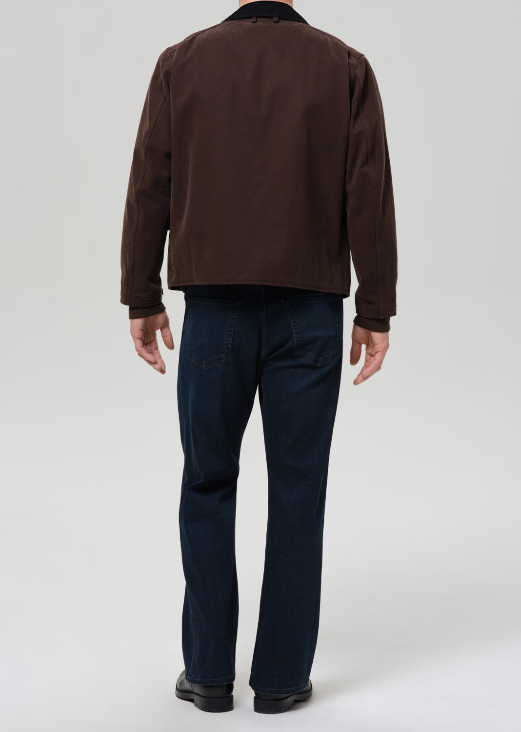 Person wearing a brown jacket and dark blue jeans on a grey background.