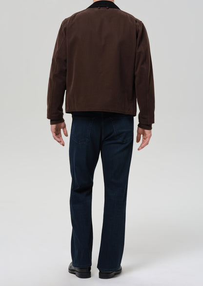 Person wearing a brown jacket and dark blue jeans on a grey background.