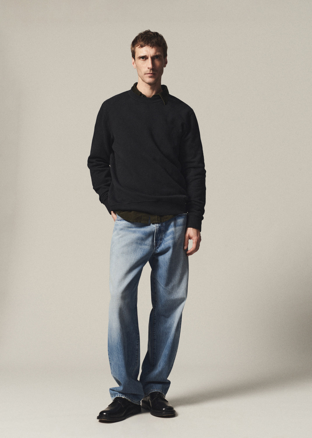 A man modeling a black long-sleeve polo shirt, light-wash blue jeans, and black leather shoes against a plain beige studio background.