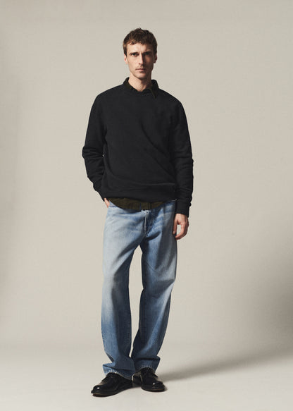 A man modeling a black long-sleeve polo shirt, light-wash blue jeans, and black leather shoes against a plain beige studio background.