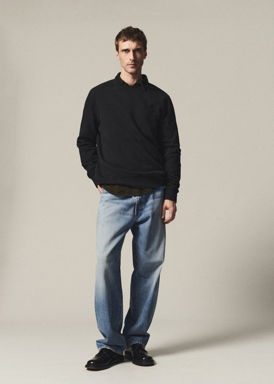 A man modeling a black long-sleeve polo shirt, light-wash blue jeans, and black leather shoes against a plain beige studio background.