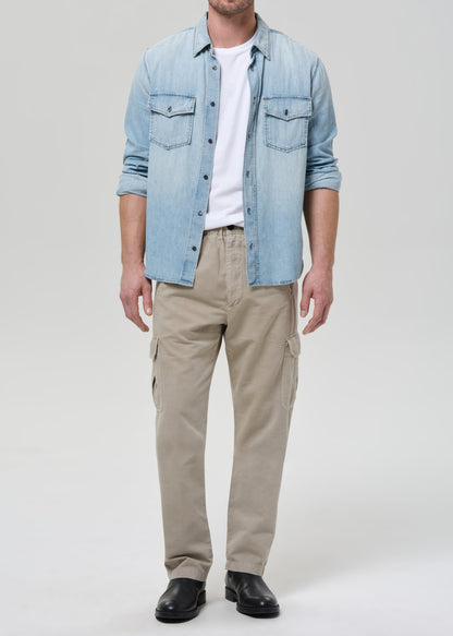 Man wearing a light blue denim shirt over a white t-shirt, beige pants, and black shoes on a plain background