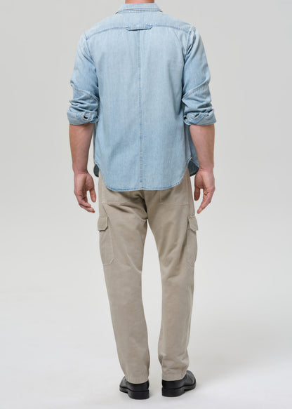Person wearing a light blue denim shirt and beige pants on a plain background