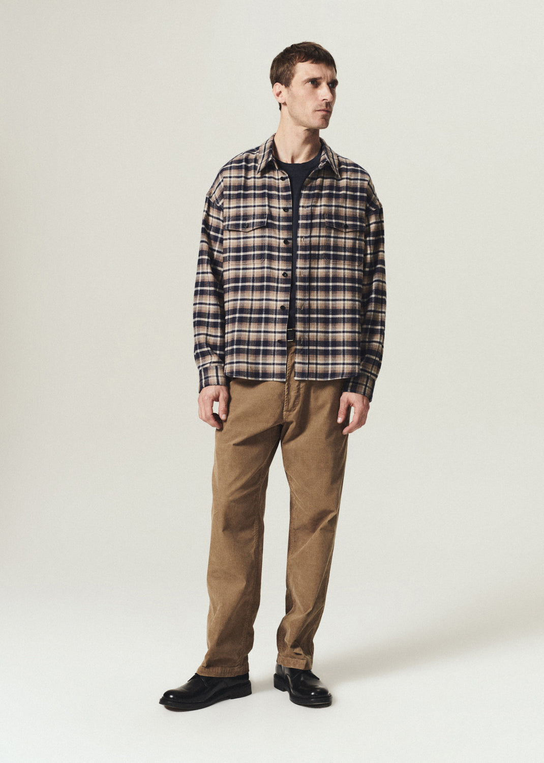 A man modeling a long-sleeve plaid button-up shirt in brown, beige, and navy, paired with brown pants and black shoes, standing against a light gray studio background.