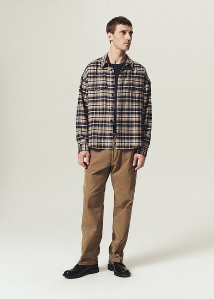 A man modeling a long-sleeve plaid button-up shirt in brown, beige, and navy, paired with brown pants and black shoes, standing against a light gray studio background.