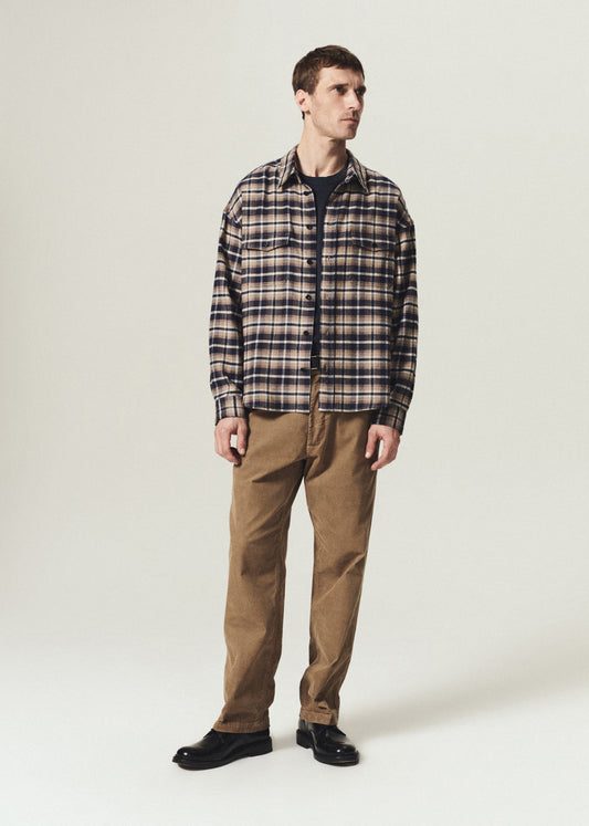 A man modeling a long-sleeve plaid button-up shirt in brown, beige, and navy, paired with brown pants and black shoes, standing against a light gray studio background.