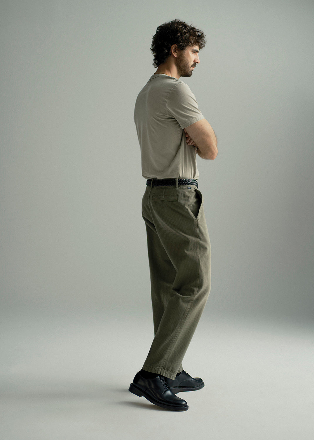 A man stands in profile wearing a light beige short-sleeve t-shirt, olive green trousers, and black leather shoes against a neutral gray studio background.