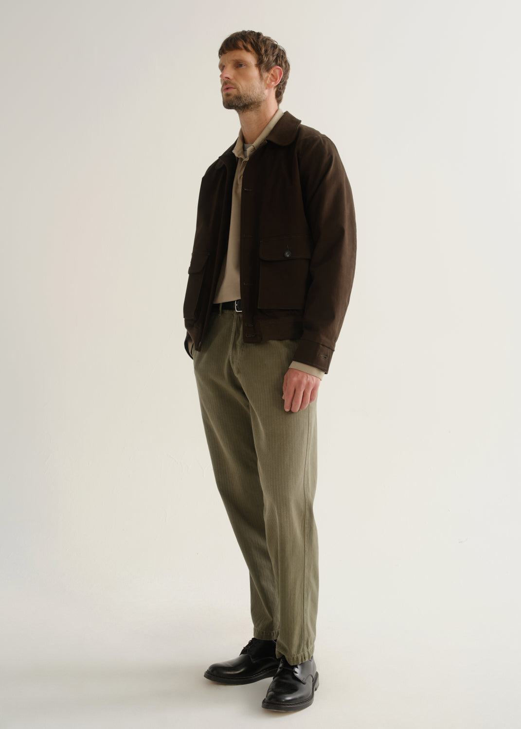 Person wearing olive green relaxed straight leg pants with a dark brown jacket and black boots, angled front full body view on a light grey background.